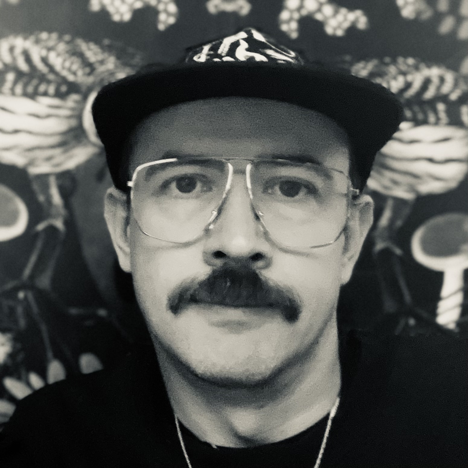 Portrait of Christopher Bennell. Black and white, wearing a hat and glasses and a gold chain. Patterned wallpaper in the background.